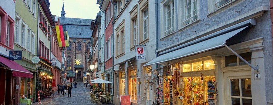 Steingasse street in Heidelberg Old Town with colorful buildings, shops, and a church in the background at dusk